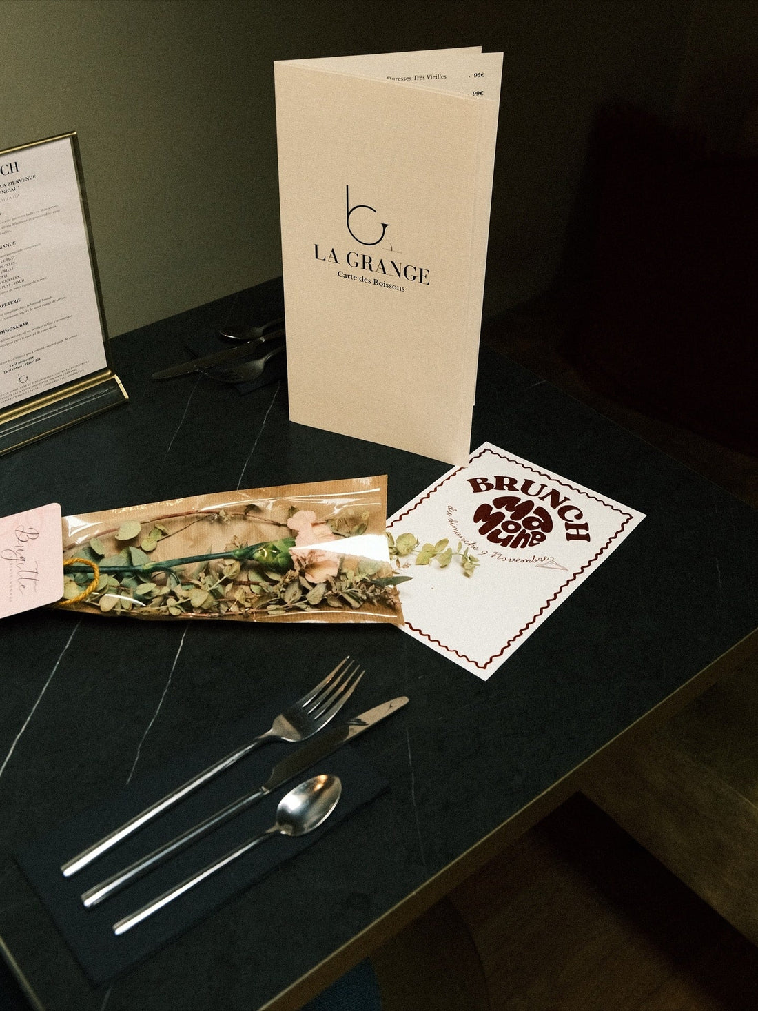 Menu du restaurant La Grange posé sur une table dressée avec des couverts, un flyer de la boutique Mamoune et un bouquet séché décoratif.